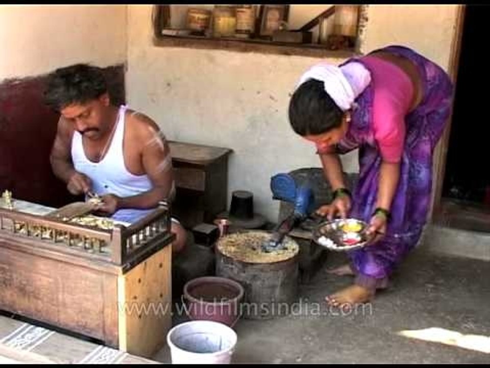 A goldsmith at work as wife performs puja