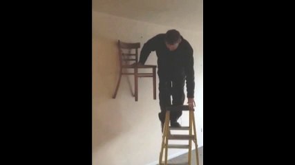 Man Sits On Chair Nailed to Wall