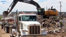 Deere 470G excavator loading debris into a double big rig dump truck on a windy day construction site