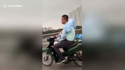 Man rides down busy road on motor scooter while holding sleeping baby