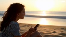 شابّة جالسة على شاطئ البحر أثناء غروب الشمس، تُمسك هاتفها بيدها وتراقب الأفق المذهل بألوانه البرتقالية والذهبية.