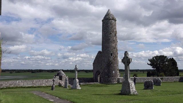 Ireland Clonmacnoise A Round Tower Stands Amid Celtic Crosses By The Shannon River