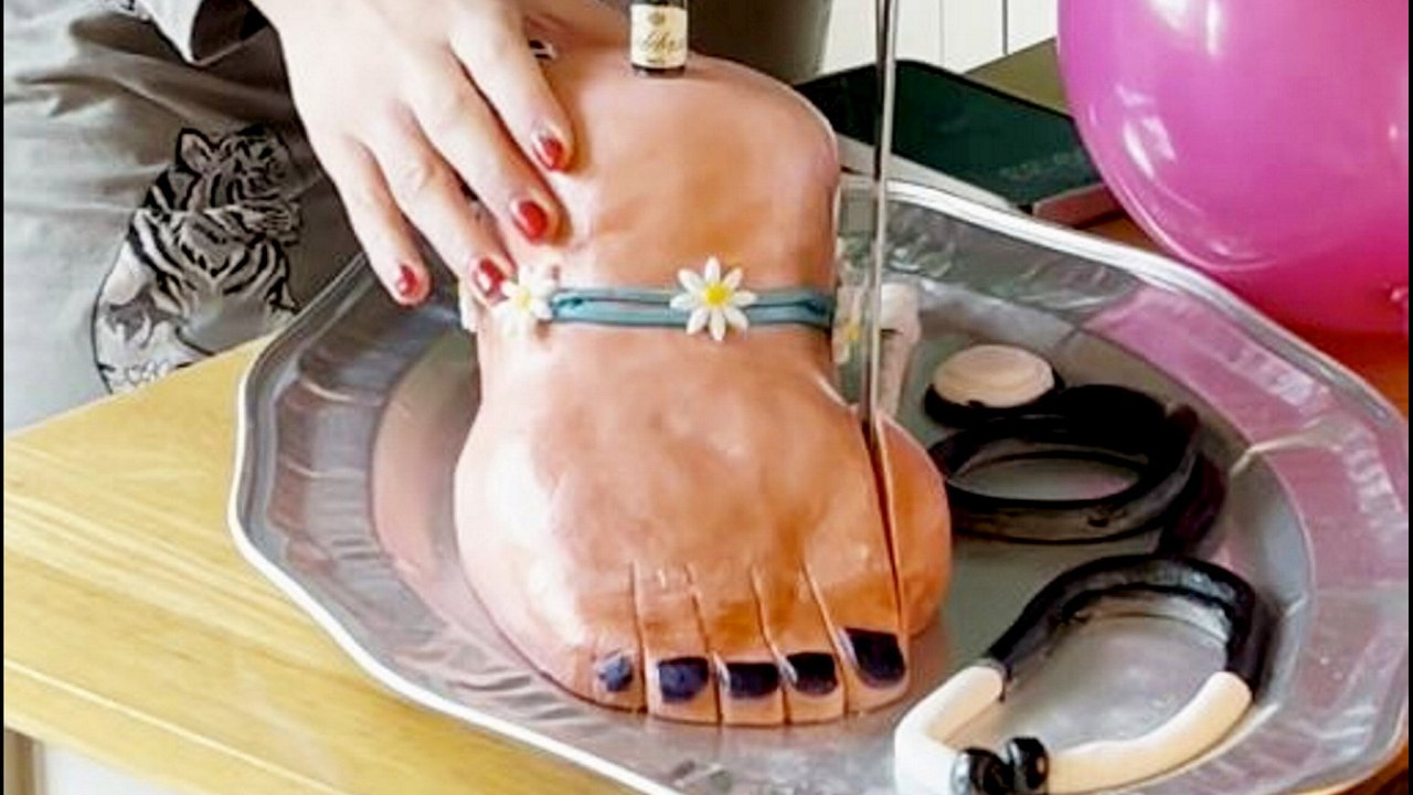 Frau feiert Geburtstag und Fußoperation, indem sie Ballenzehe von fußförmigem Kuchen abschneidet