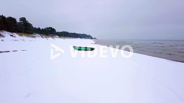 Aerial establishing view of Baltic sea coast on a overcast winter day with green coastal fisherman boat, beach with white sand covered by snow, coastal erosion, wide angle drone shot moving forward
