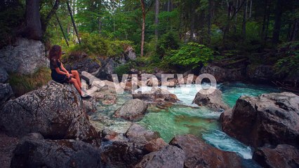 4K UHD Cinemagraph - seamless video loop of a young brunette woman sitting at a river at the scenic mountain lake Hintersee in Bavaria, Germany
