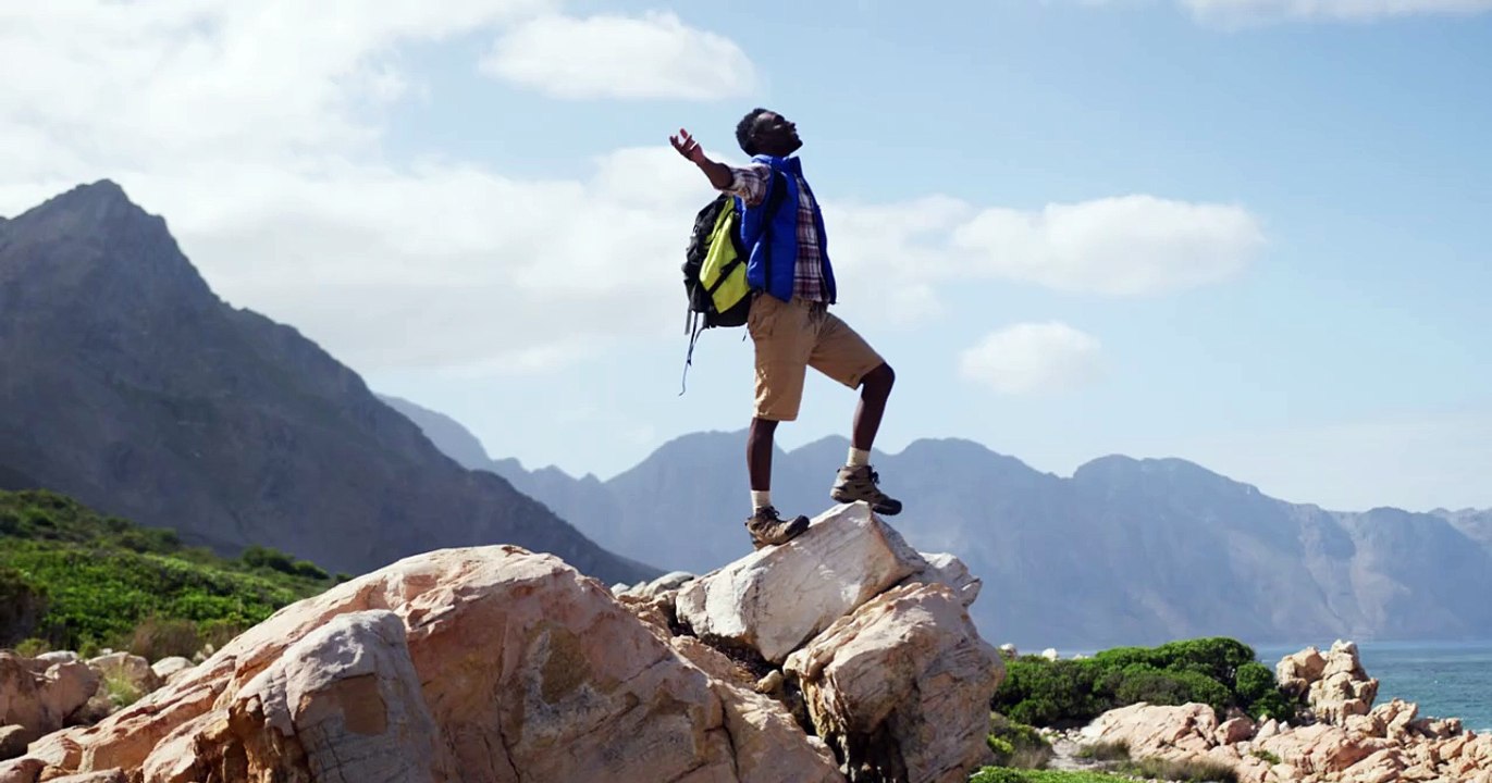 african american man standing on rock with arms wide open while trekking in the mountains