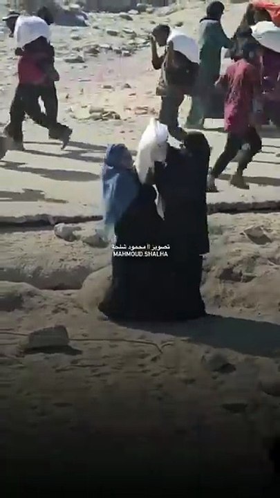 Two Palestinian women take turns carrying a bag of flour on their exhausted shoulders, walking with heavy steps on a long, dangerous road, after obtaining it from the heart of the "death traps" northwest of Gaza.
