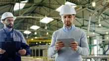 caucasian male worker wearing a helmet and using a tablet in a factory, then his coworker comes up and talking to him