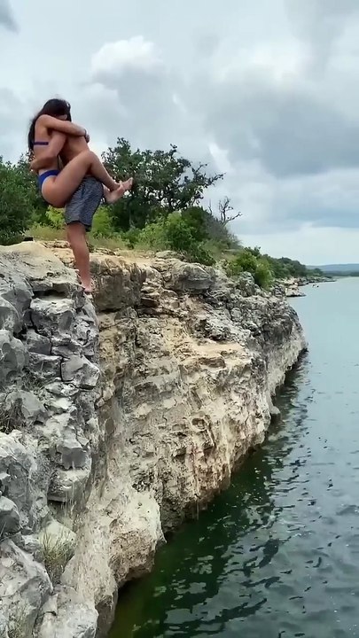 This image shows a person jumping off a rocky cliff into the water while doing a flip in mid-air. The surroundings include rugged rocks, some greenery and trees in the background, and clear water below. The moment is captured right in motion, reflecting a